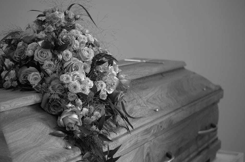 A large floral arrangement of mixed roses and other flowers in various stages of bloom, with delicate petals and lush greenery, rests on the top of a closed wooden coffin with a rounded lid. The bouqu