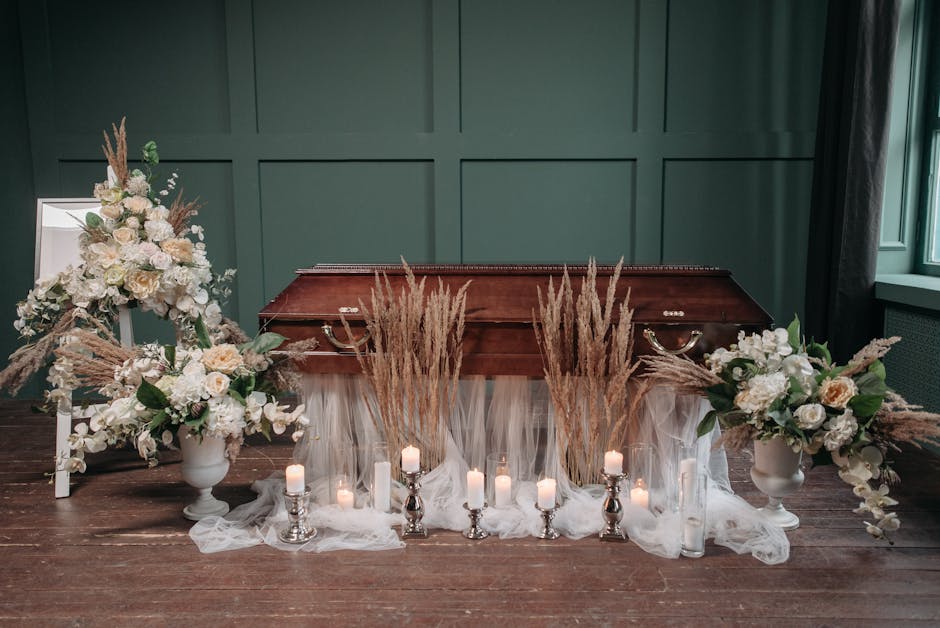 A funeral floral arrangement featuring two large white vases filled with white roses, hydrangeas, and eucalyptus leaves, flanking a classical wooden coffin with a dark finish. Behind the coffin, tall 