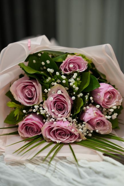 A bouquet of fresh, pale pink roses with tightly layered petals, accented by small white baby's breath flowers and lush green foliage. The flowers are wrapped in elegant white tissue paper with a soft